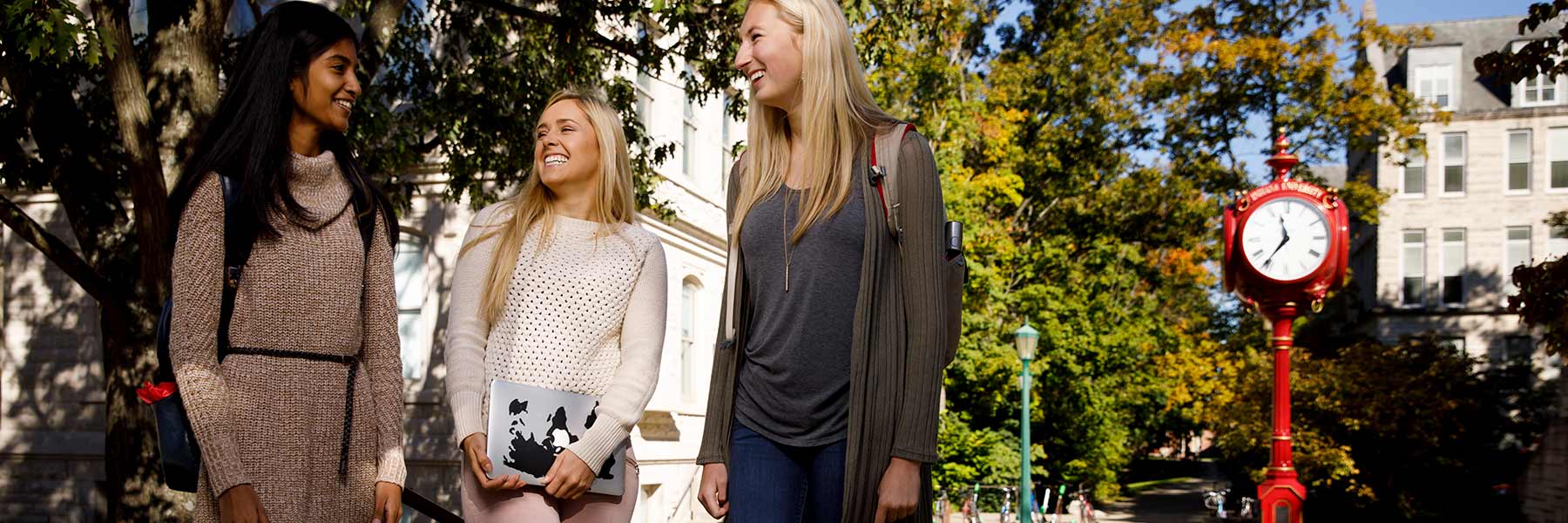 Three students stand together and laugh outside of Maxwell Hall near an IU red clock on campus.